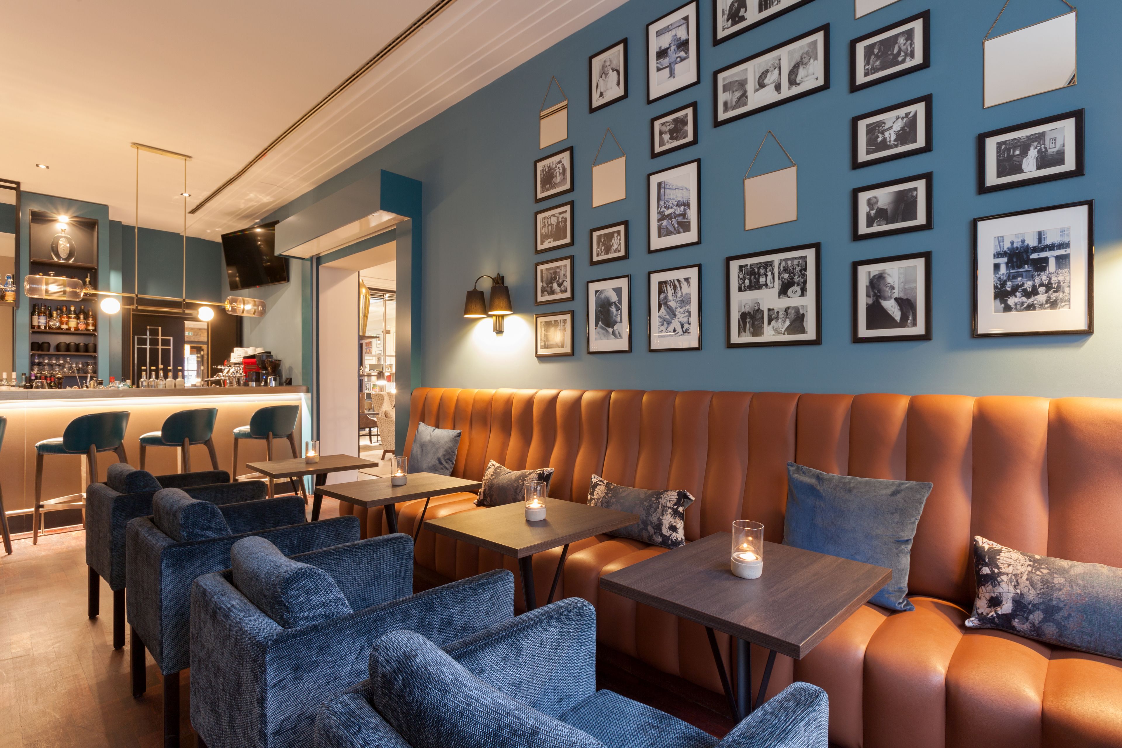 Un rincón de bar acogedor con asientos de cuero, sillas tapizadas en azul y una pared decorada con fotografías enmarcadas.