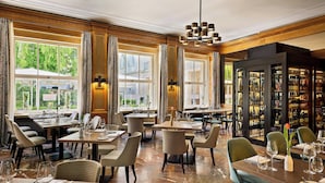 A restaurant with multiple dining tables and chairs, large windows with curtains, and a wine display cabinet. - Hotel Elephant Weimar, Autograph Collection (Weimar)
