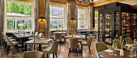 A restaurant with multiple dining tables and chairs, large windows with curtains, and a wine display cabinet.
