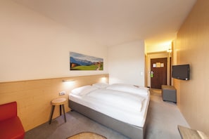 Una habitación de hotel con una cama, un taburete pequeño, un televisor y un cuadro en la pared.