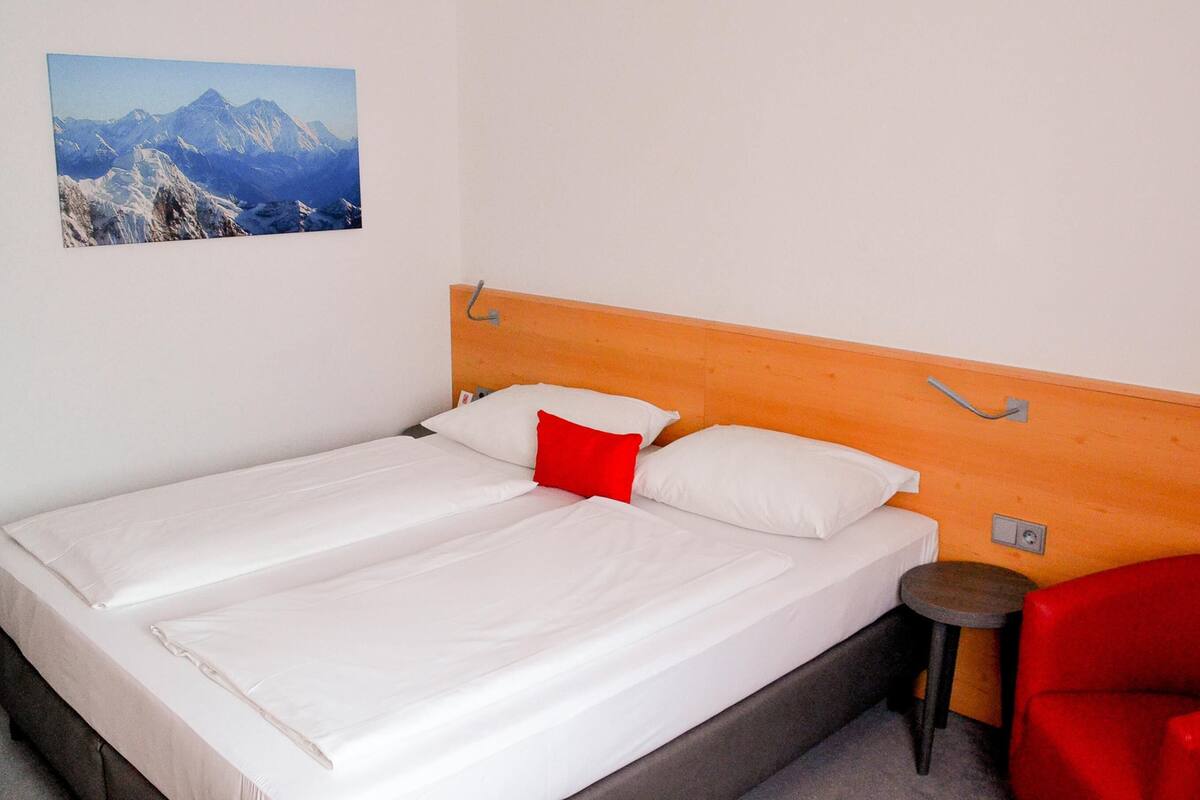 A bed with white bedding and a red pillow, a wooden headboard, a small stool, and a red chair.