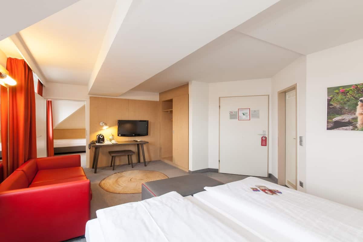 A hotel room with a red sofa, a bed, a desk with a chair, a television, and a door with a fire alarm.