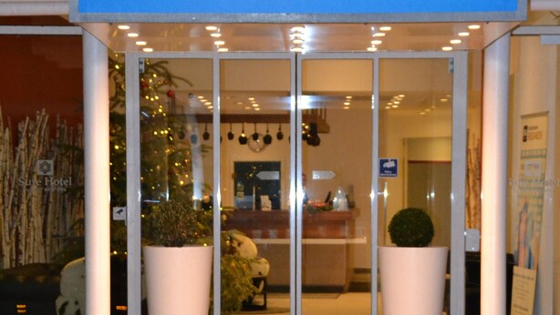 The entrance of Sure Hotel by BEST WESTERN with a blue awning, glass doors, and potted plants.