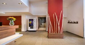 A hotel reception area with a wooden reception desk, a vase of flowers, a self-service kiosk, and a decorative plant arrangement. - Sure Hotel by Best Western Muenchen Hauptbahnhof (Munich)