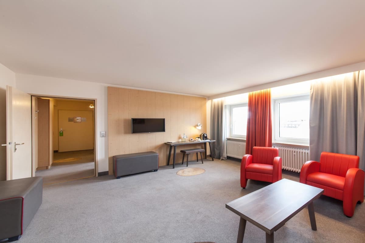 A modern hotel room with a flat-screen TV, a small table, two red sofas, a wooden bench, and a large window with curtains.