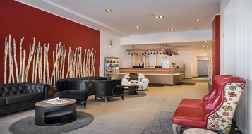 Et hotellobby med reception, siddeområde med sofaer og lænestole, en dekorativ væg med grene og et køkken i baggrunden.