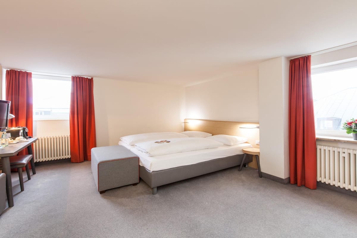 A hotel room with a bed, a desk with a chair, a television, and a window with curtains.