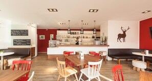 Interior de restaurante con barra, mesas de madera, sillas rojas y un vinilo decorativo de un ciervo en la pared.