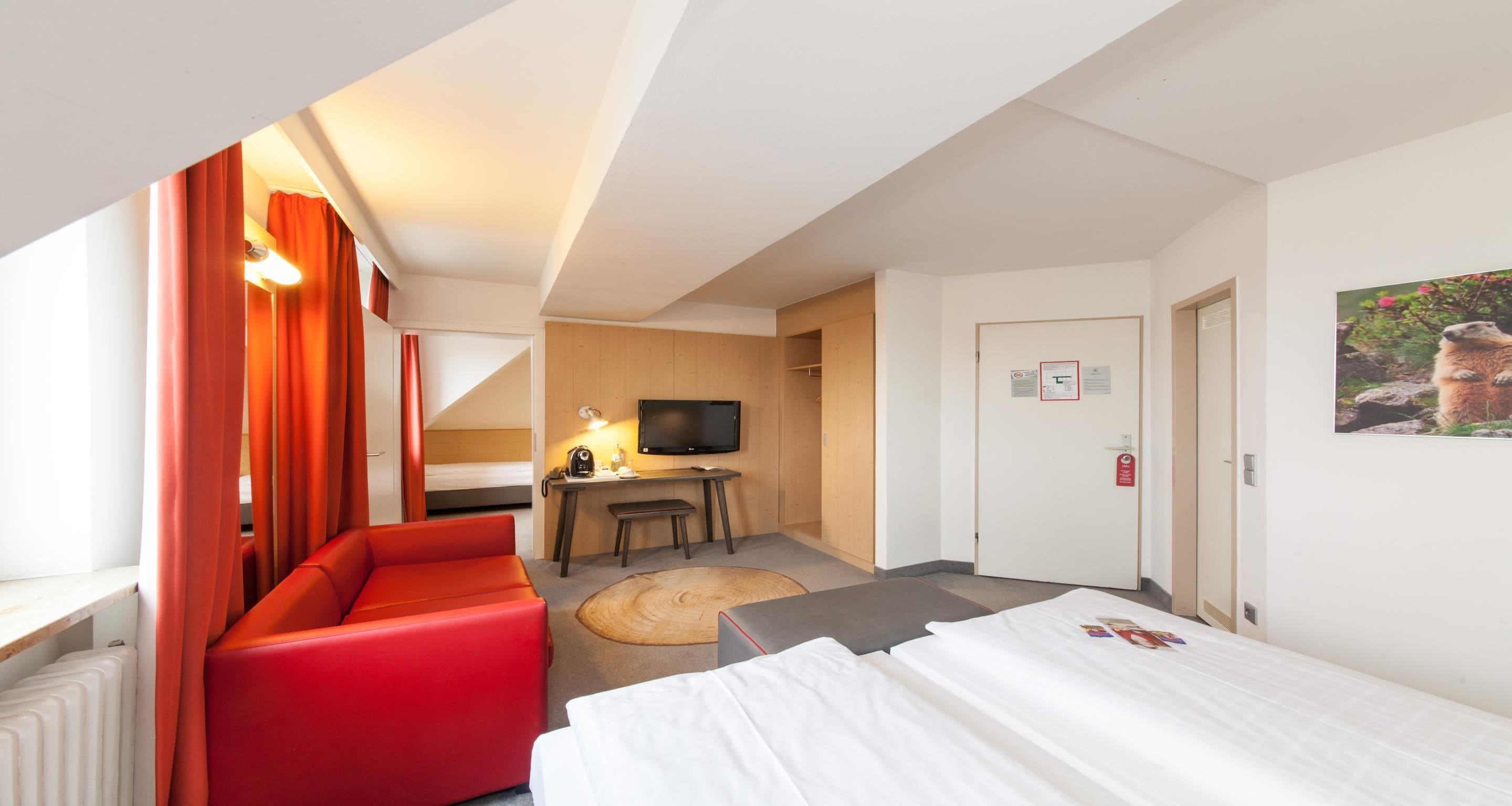 A hotel room with a red sofa, a bed, a desk with a computer, a television, and a door with a fire alarm.