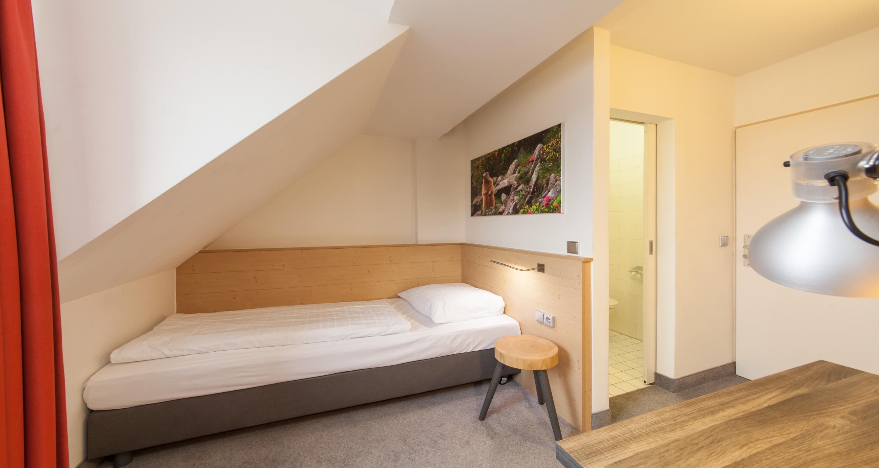 Una habitación pequeña con cama individual, techo inclinado, cabecera de madera, mesita de noche, un taburete pequeño y un cuadro en la pared.