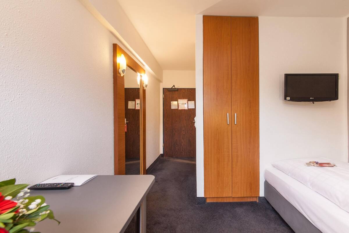 A hotel room with a bed, a TV mounted on the wall, a desk with a remote control, and a wardrobe.