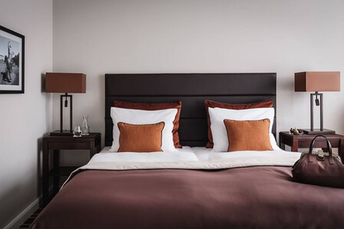 Een netjes opgemaakt bed met een donker hoofdbord, twee nachtkastjes met lampen en een ingelijst schilderij aan de muur.