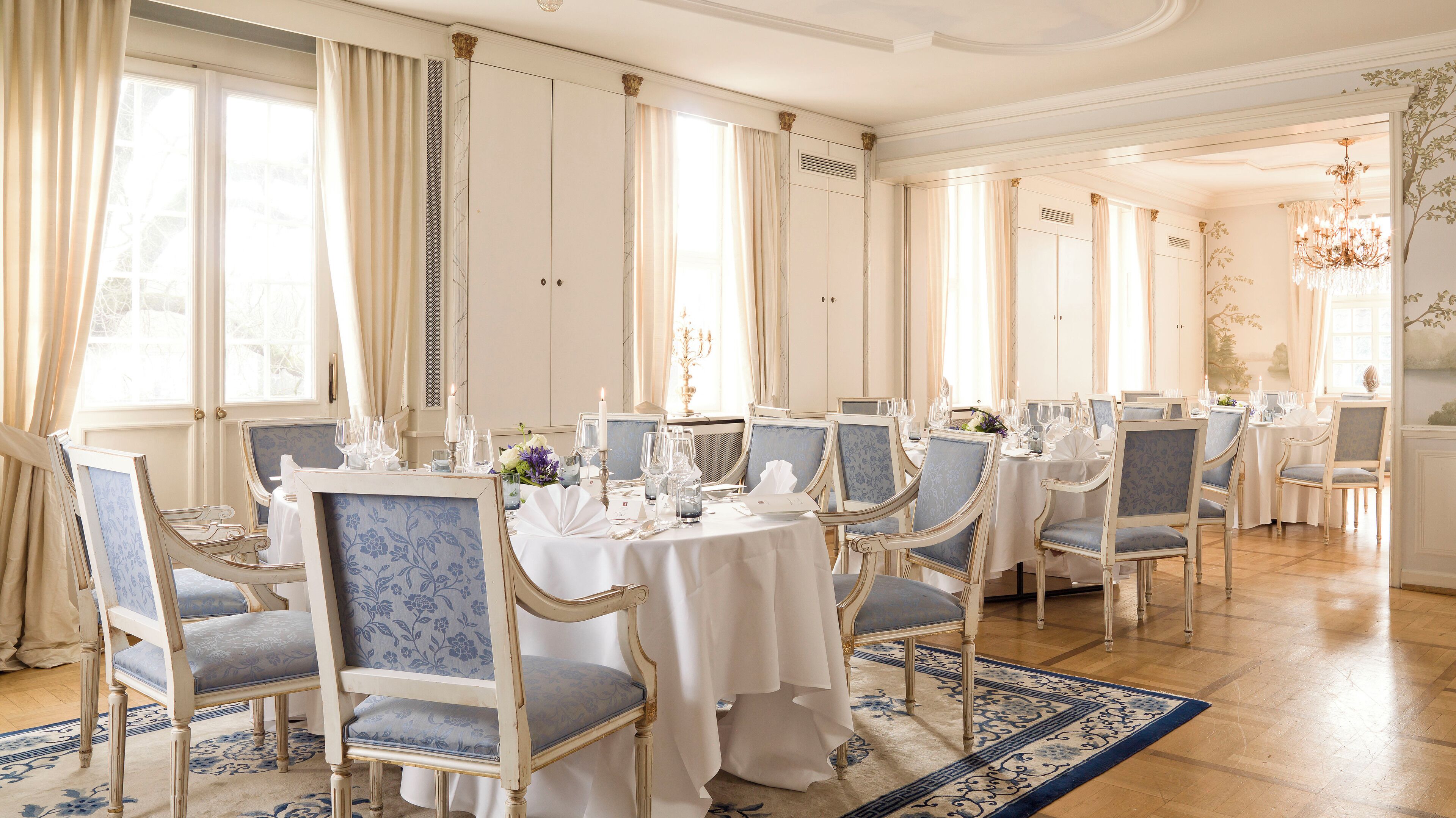 A formal dining room with multiple tables set for service, elegant chairs, and a chandelier.