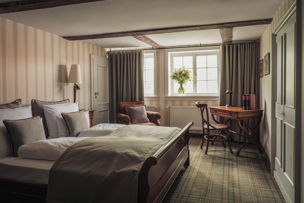 Een hotelkamer met een groot bed, een bureau en een stoel. Er is een raam met gordijnen en een bloemenvaas op de vensterbank.