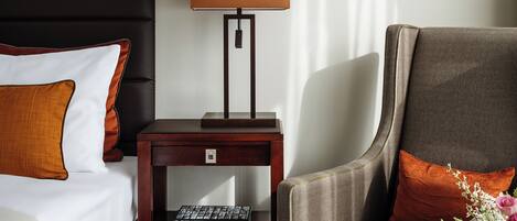 A hotel room with a bed, a desk with a lamp, a chair, and a small table with a vase of flowers.