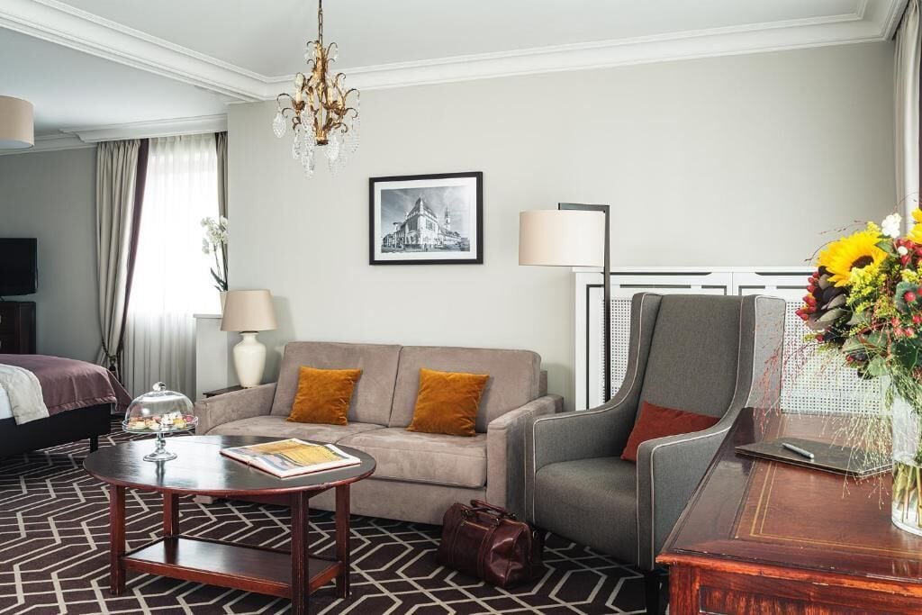 Habitación de hotel con cama, sofá, sillón, mesa, lámpara, un cuadro en la pared y un florero con flores.