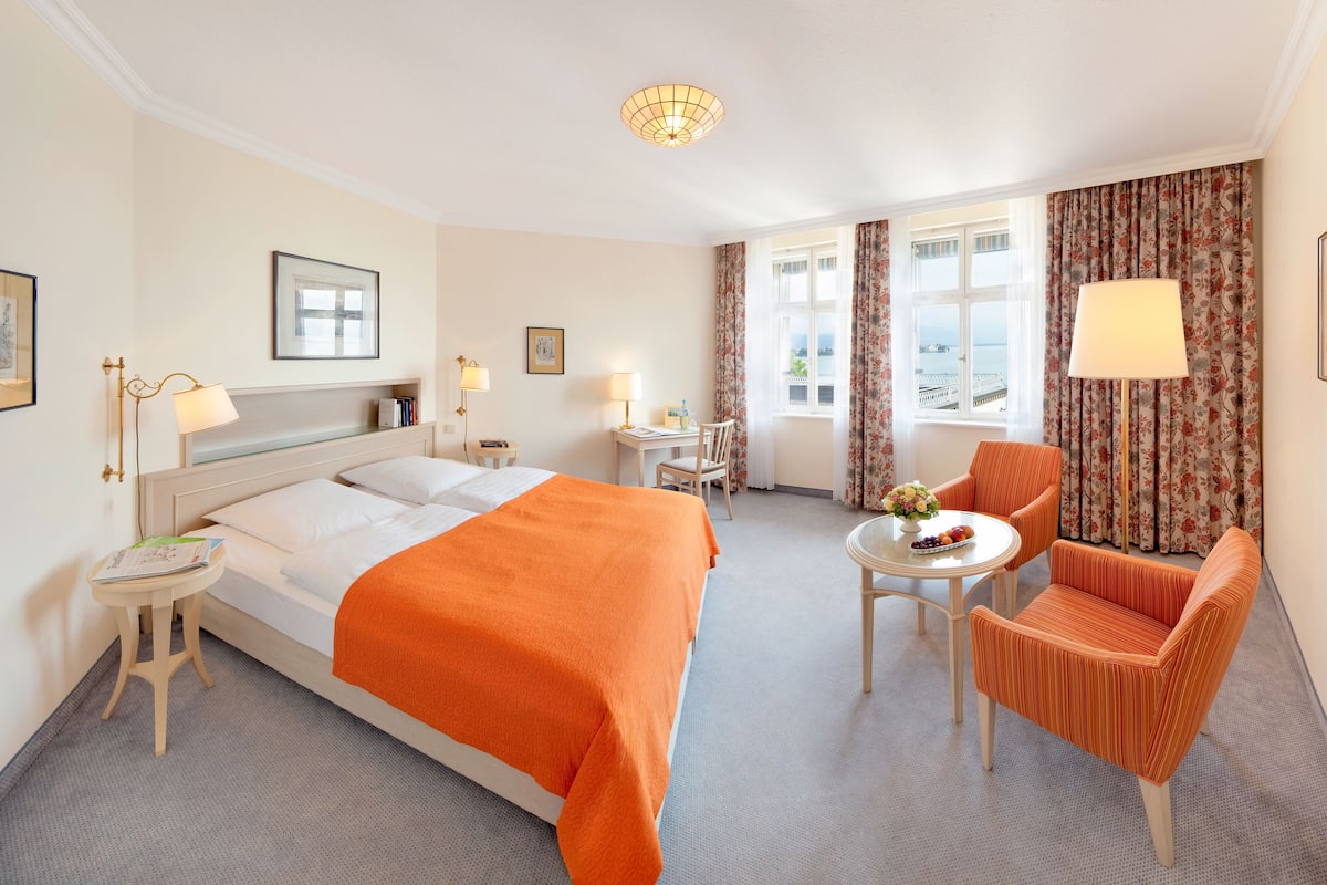 A hotel room with a bed, a chair, a table with fruit, and a lamp.