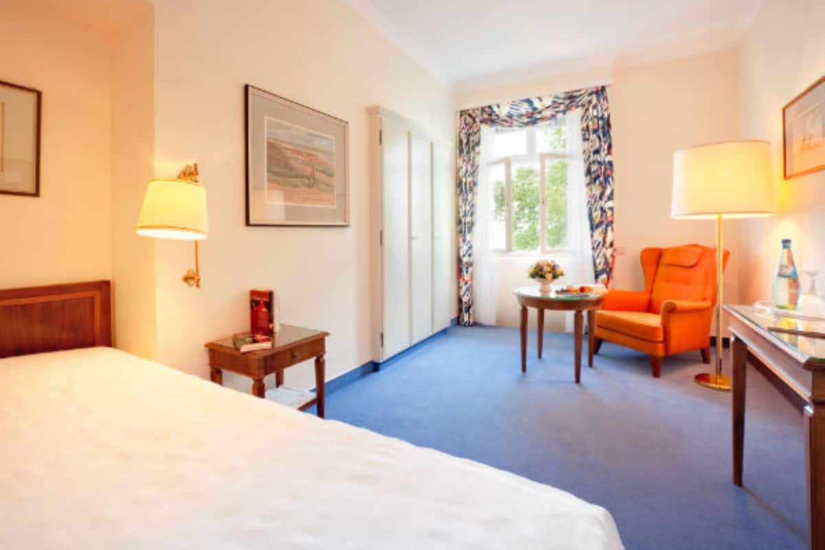 A hotel room with a large bed, a desk with a lamp, an orange armchair, and a window with patterned curtains.