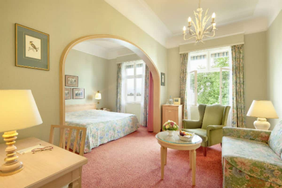 A hotel room with a bed, two chairs, a small table, a desk, a lamp, a mirror, and a window with curtains.