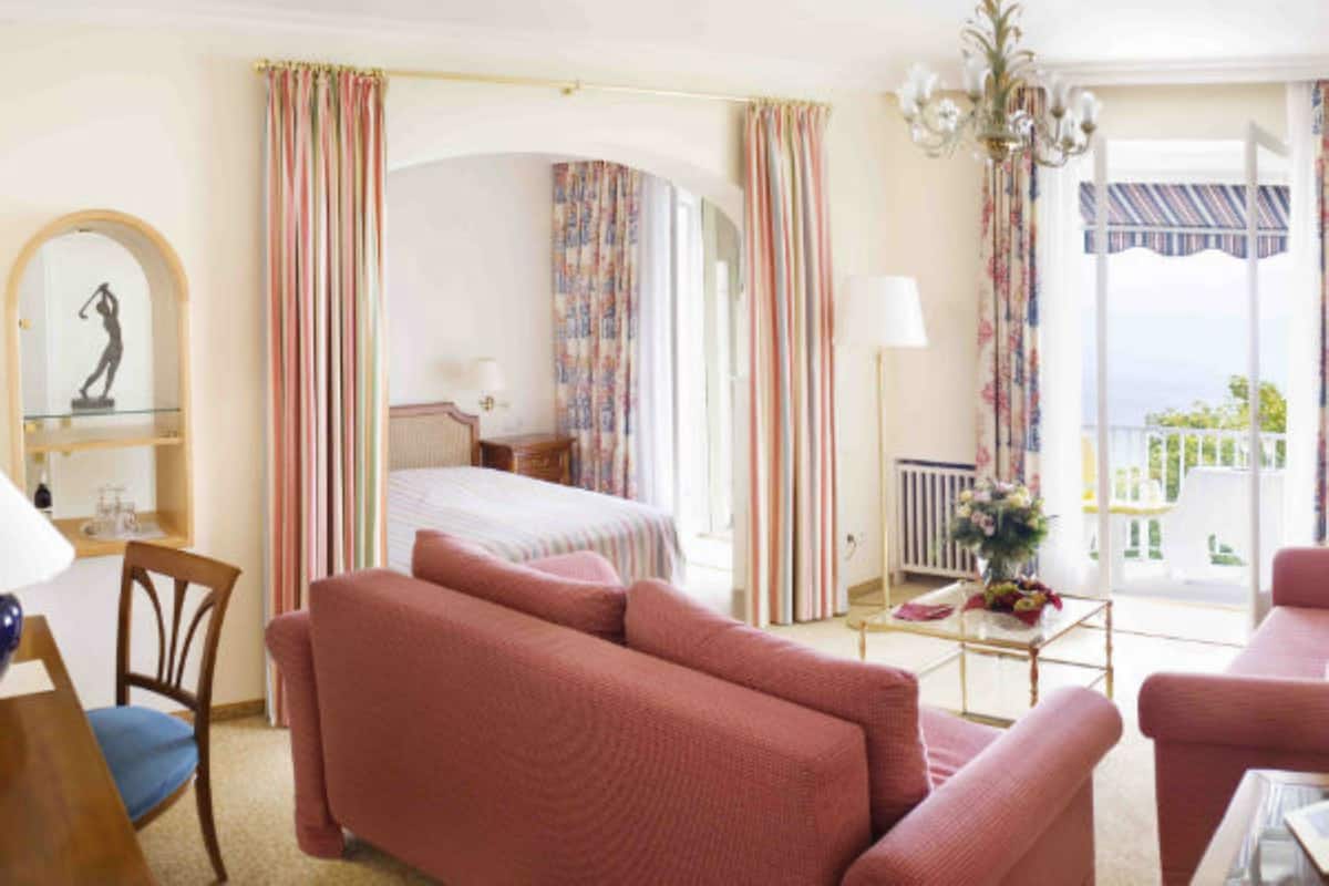 A hotel room with a bed, a sitting area with a sofa and armchairs, a desk with a chair, a chandelier, and a view of the outdoors.
