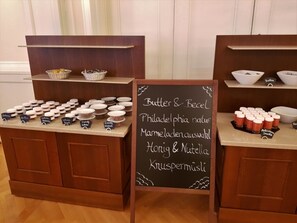 Un buffet proposant différents plats présentés dans des récipients étiquetés, ainsi qu’un tableau noir affichant le menu avec des options telles que « Beurre et miel », « Philadelphia nature », « Assortiment de confitures », « Miel et Nutella » et « Muesli aux noix ».