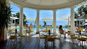 Un espacio de estar techado al aire libre con mesas y sillas, con vistas a una piscina y al mar.
