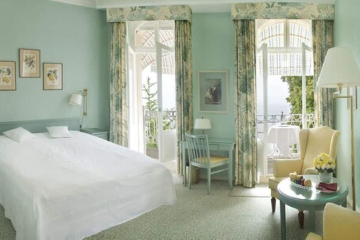 A hotel room with a large bed, a chair, a small table with flowers, and a balcony with a view.
