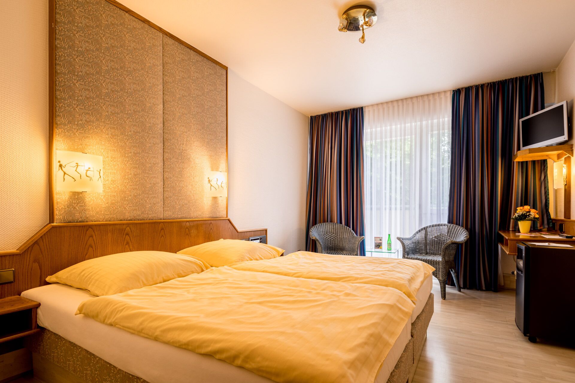 Una habitación de hotel con una cama grande, dos sillas, un escritorio con televisor y un ventanal con cortinas.