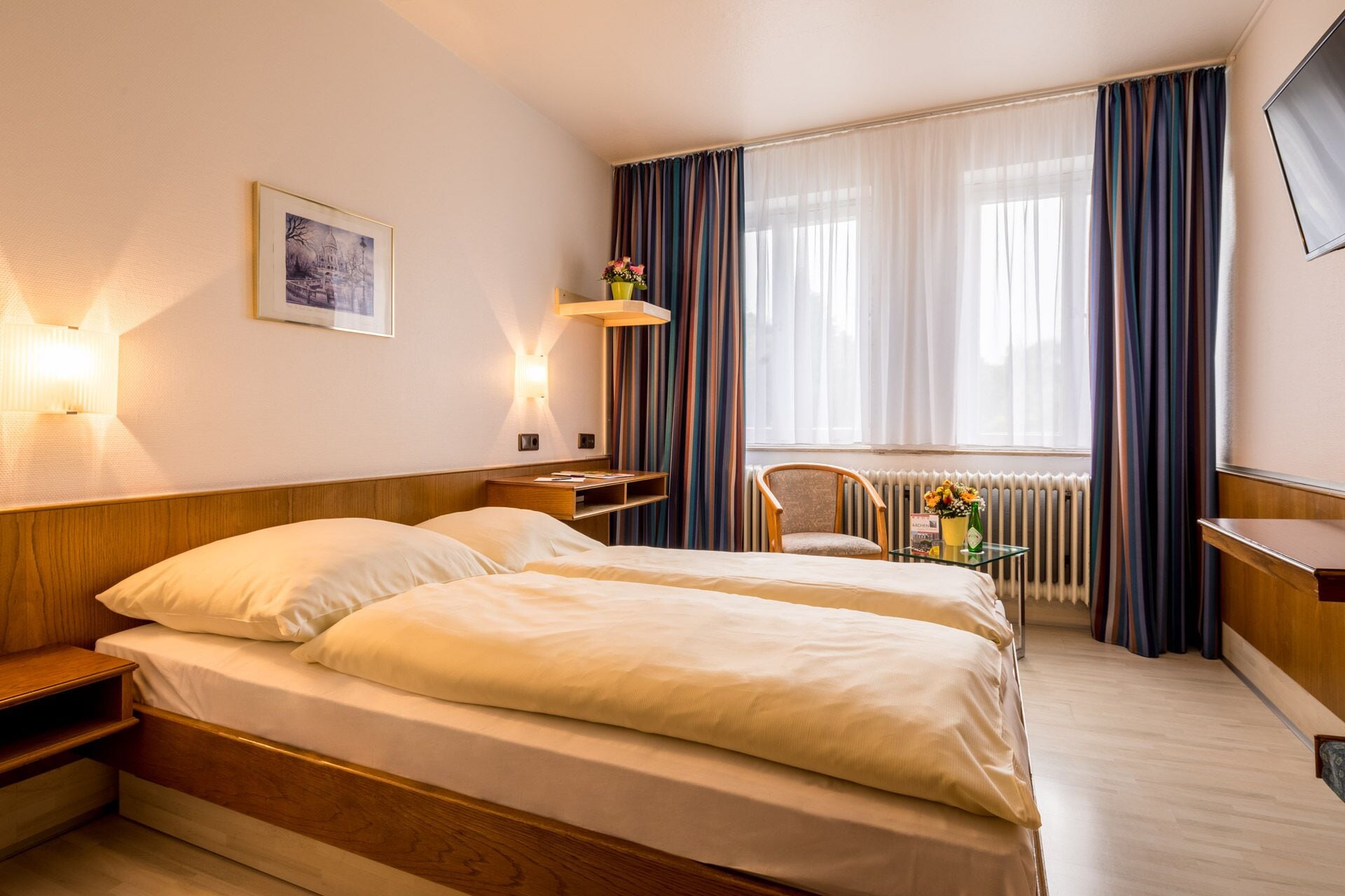 Ein Hotelzimmer mit zwei Betten, einem Schreibtisch, einem Sessel, einem Heizkörper und einem Fernseher.