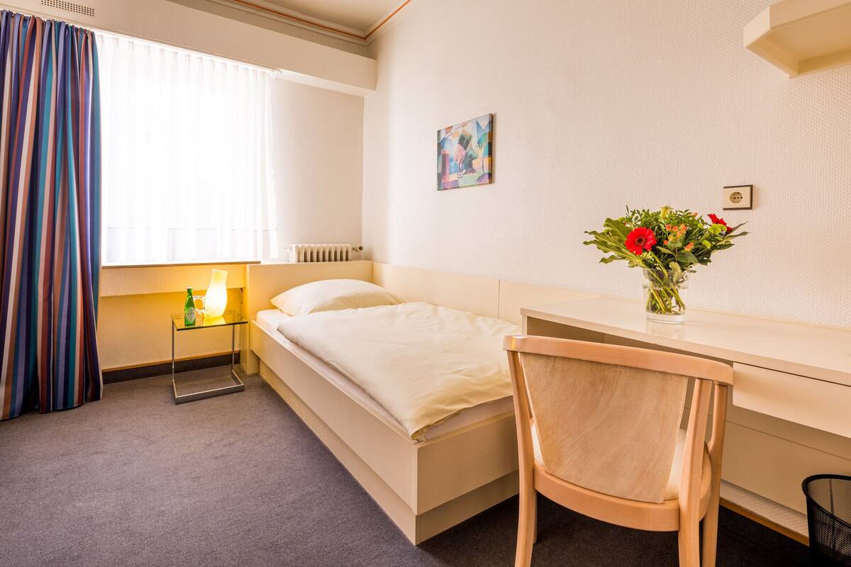 Habitación de hotel con cama, escritorio, silla, lámpara, radiador, un cuadro y un jarrón con flores.