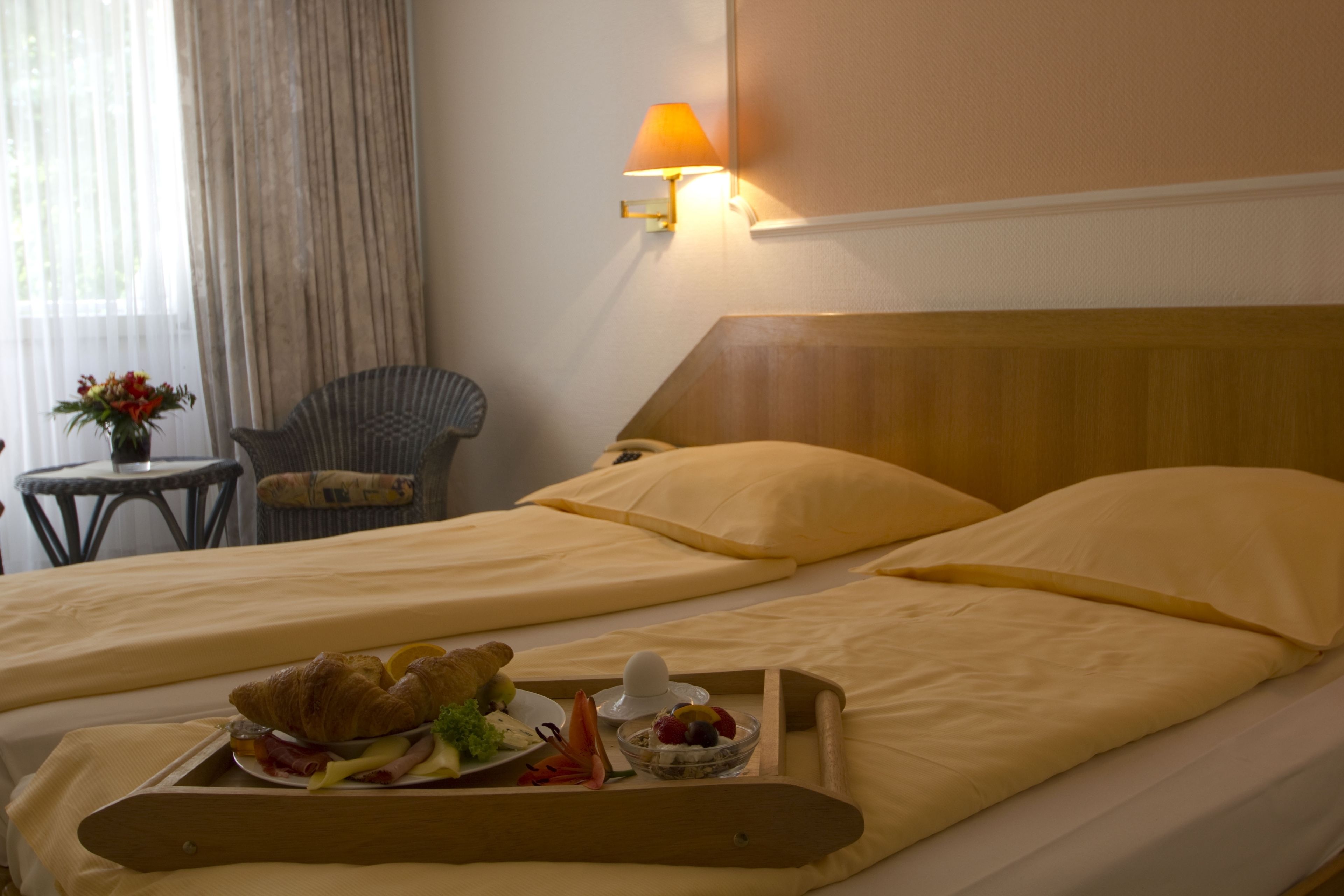 A hotel room with two beds, a bedside table with a lamp, a chair, and a tray with breakfast items.