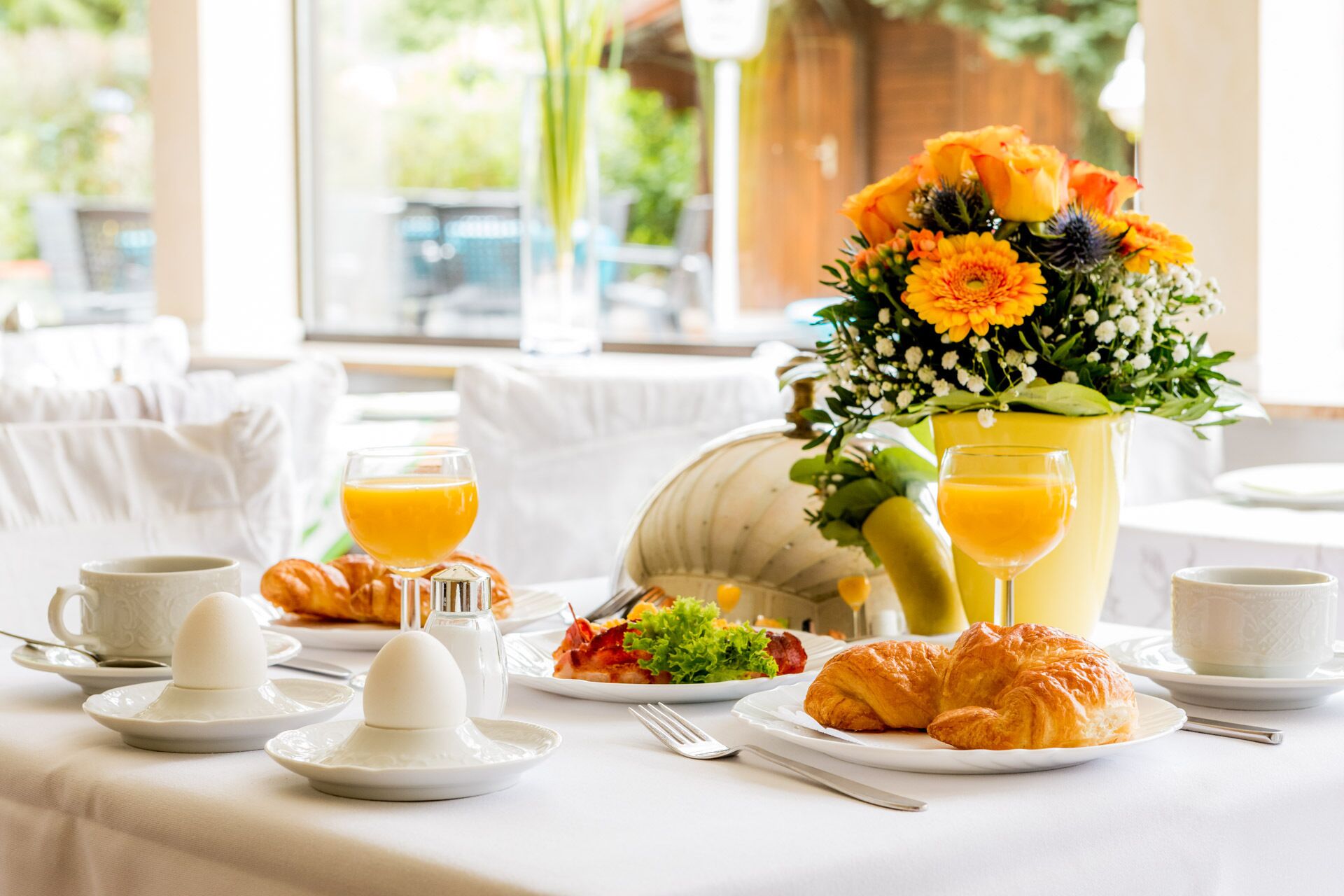 Un desayuno con croissants, huevos, jugo de naranja y un arreglo floral.