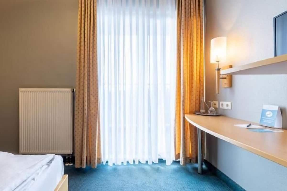 A hotel room with a bed, a desk with a lamp, a radiator, and a window with curtains.