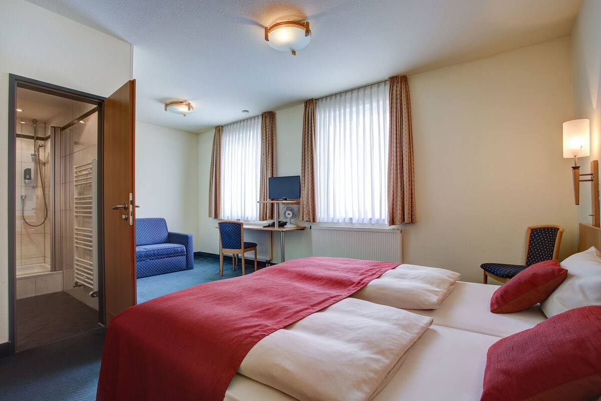 A hotel room with a bed, a desk with a computer, a chair, a TV, and a shower area.