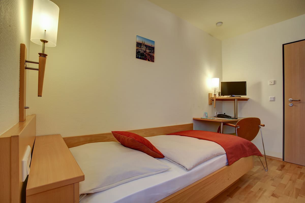 A single bed with a wooden frame, a bedside table, a lamp, a desk with a chair, and a TV.