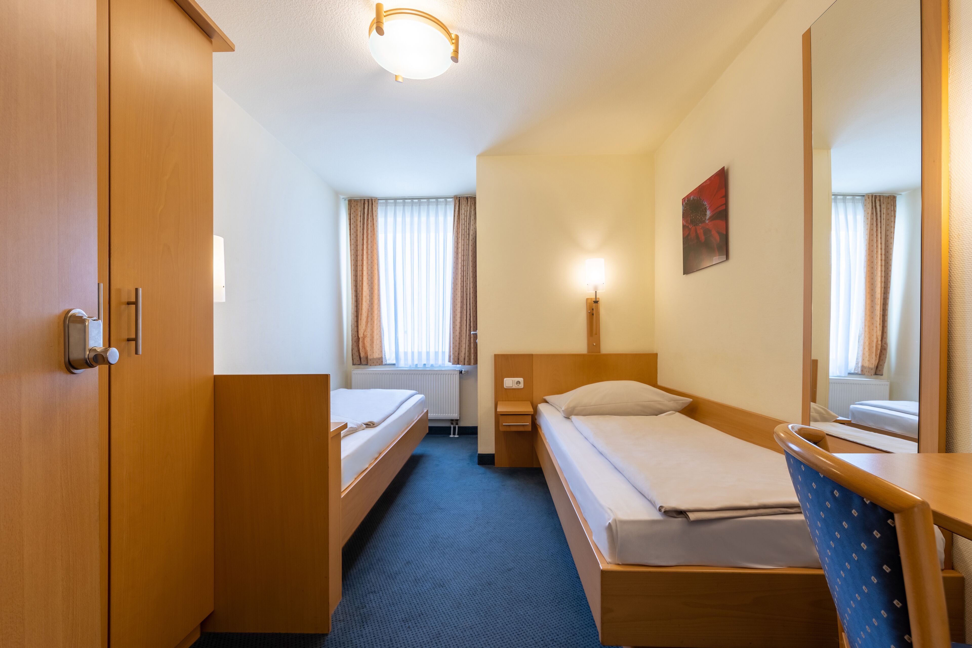 A hotel room with a single bed, a desk, a chair, a wardrobe, and a window with curtains.