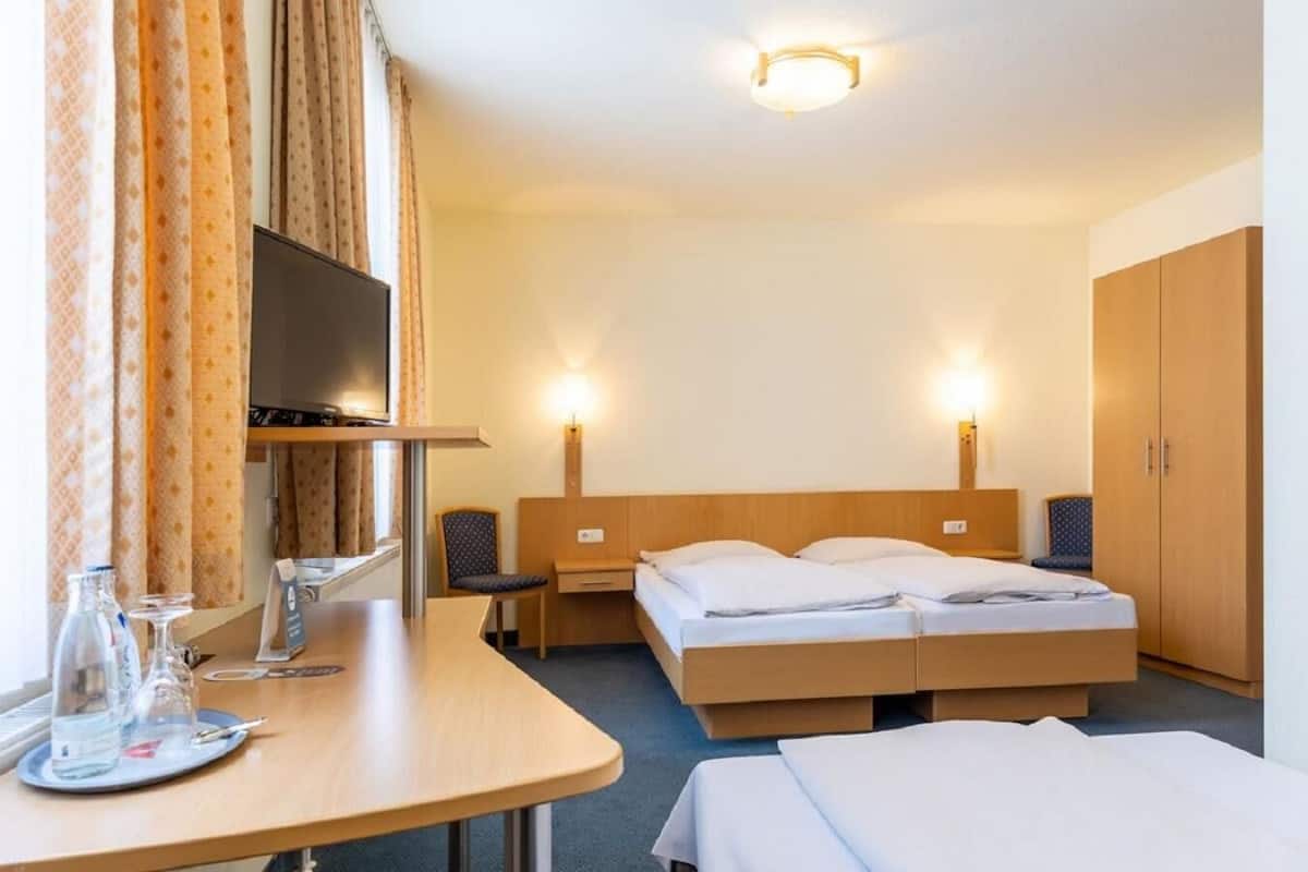 A hotel room with a bed, bedside tables, a TV, and a desk with glasses and a bottle.