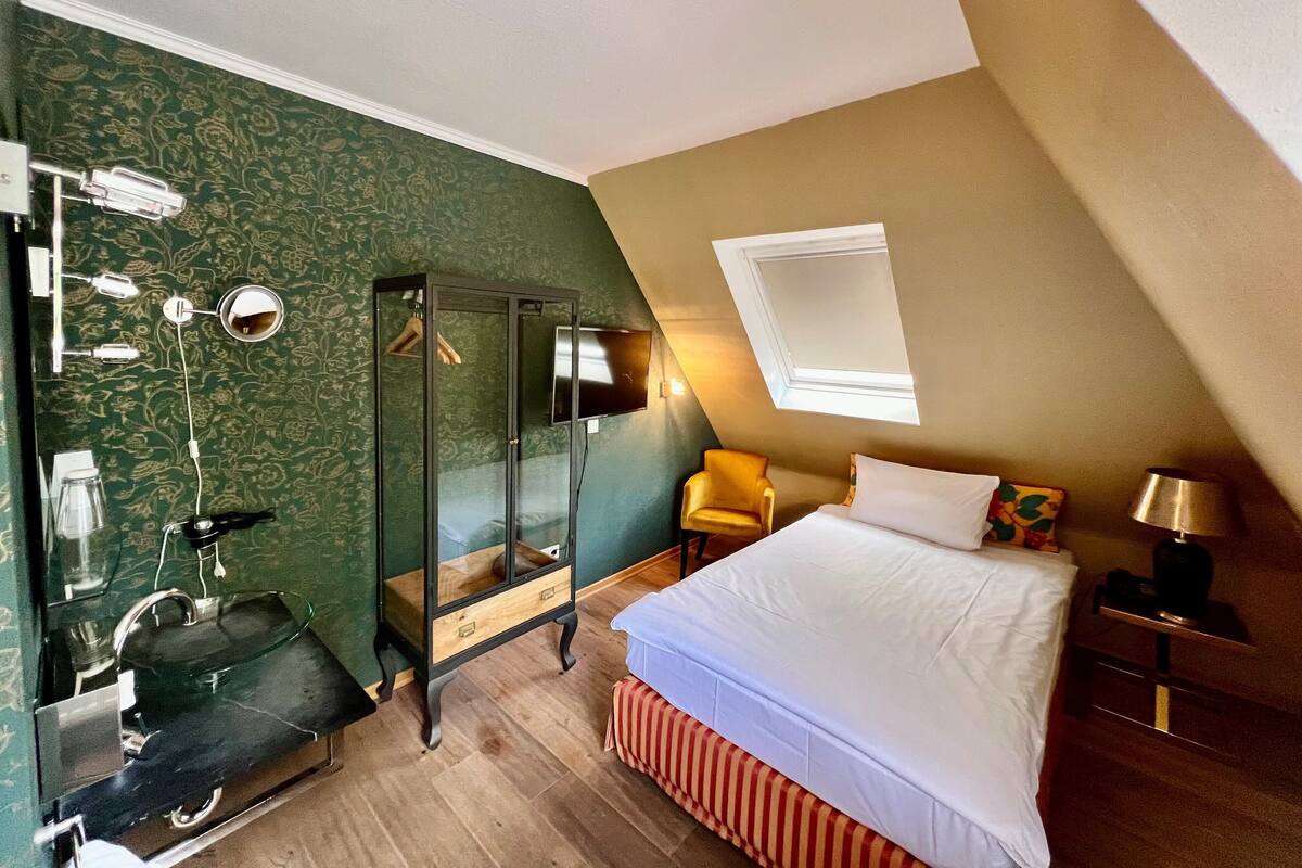 A bedroom with a bed, a bedside table, a lamp, a chair, a mirror, and a shower area.