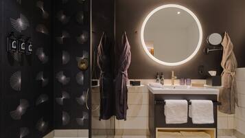 Un baño moderno con una pared negra que presenta un patrón de luz único, un espejo redondo con un halo luminoso, un tocador con un lavabo y dos toallas, y una cabina de ducha con paredes de cristal.