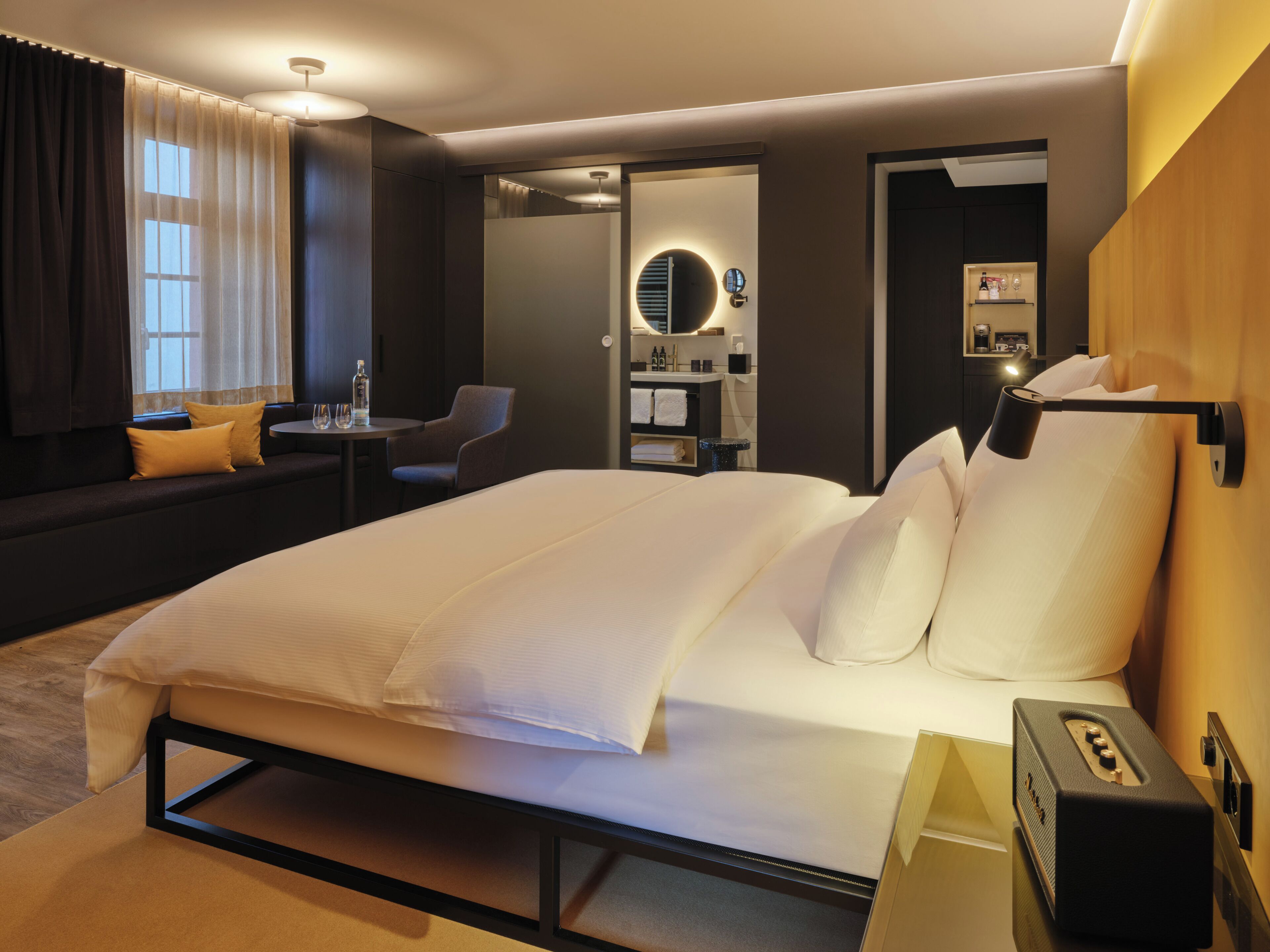 Et moderne hotelværelse med en stor seng, et siddeområde med et bord og stole, et sminkebord med spejl og en lille hylde med toiletartikler.