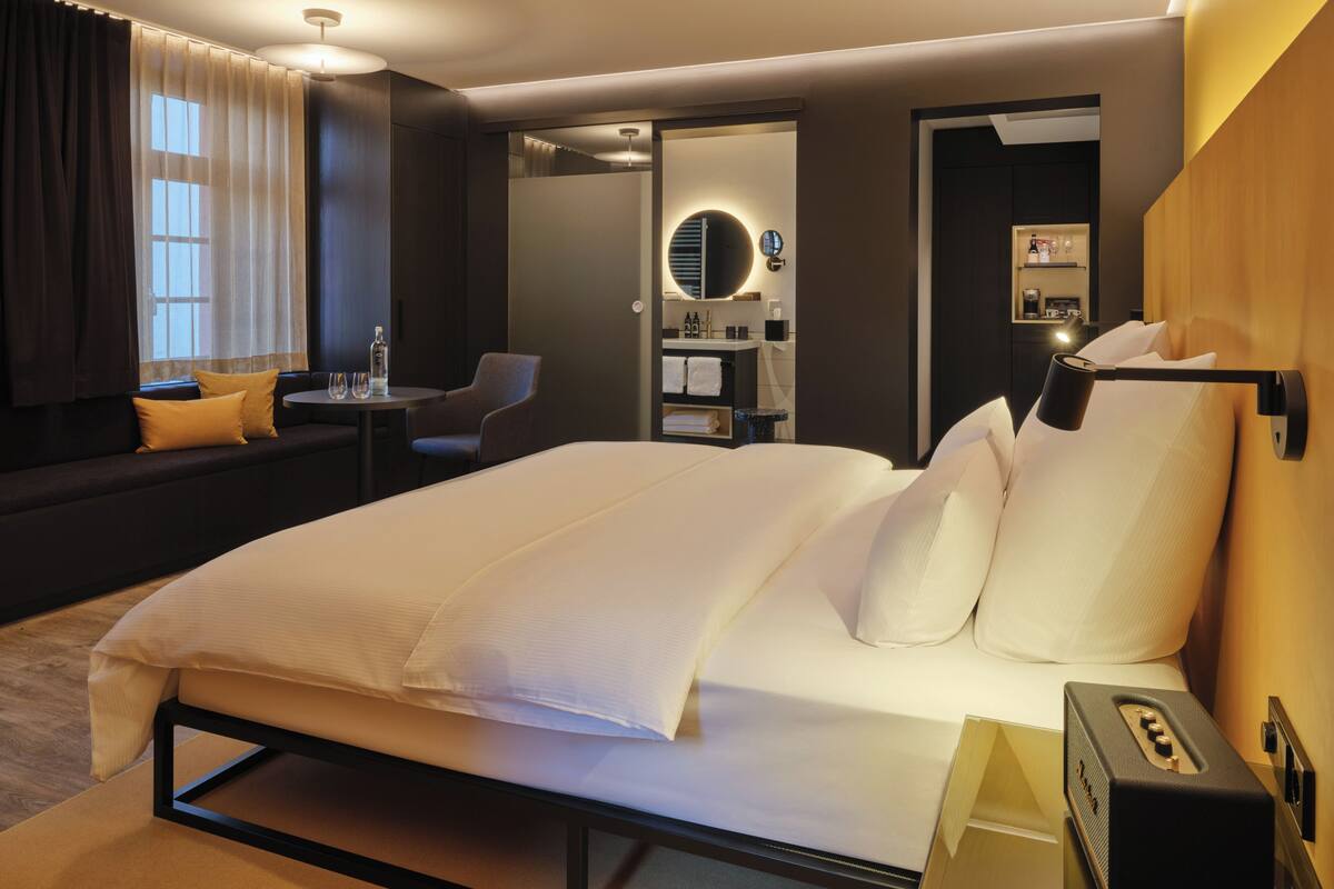 Une chambre d’hôtel moderne dotée d’un grand lit, d’un coin salon avec une table et des chaises, d’une coiffeuse avec un miroir et d’une petite étagère contenant des articles de toilette.
