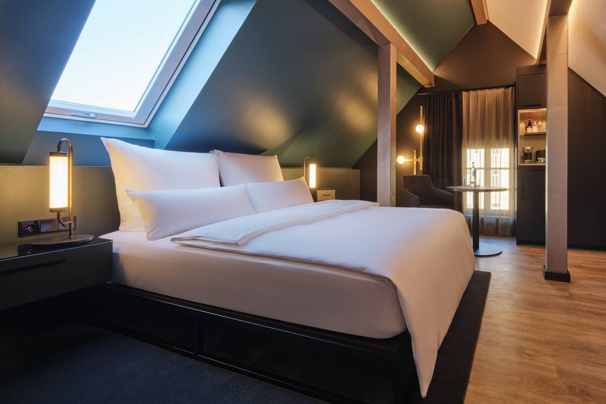 Une chambre d’hôtel moderne avec une lucarne, un grand lit, des lampes de chevet, une petite table et une chaise.