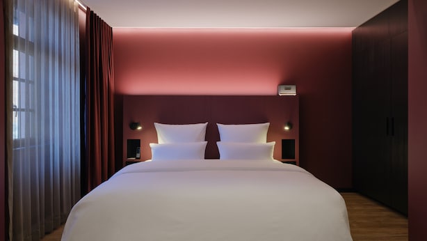 Un grand lit recouvert de draps blancs, dans une pièce aux murs rouges et au plancher en bois.
