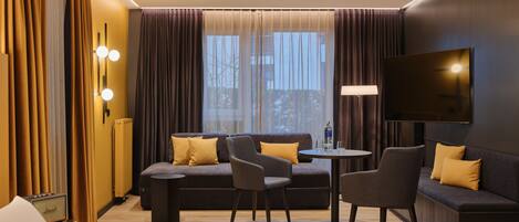 A modern hotel room with a sofa, two armchairs, a small table, and a flat-screen TV mounted on the wall.