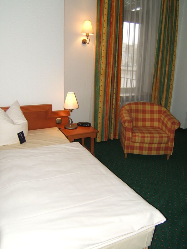 Una habitación de hotel con cama, mesita de noche con lámpara, una silla y una ventana con cortinas.