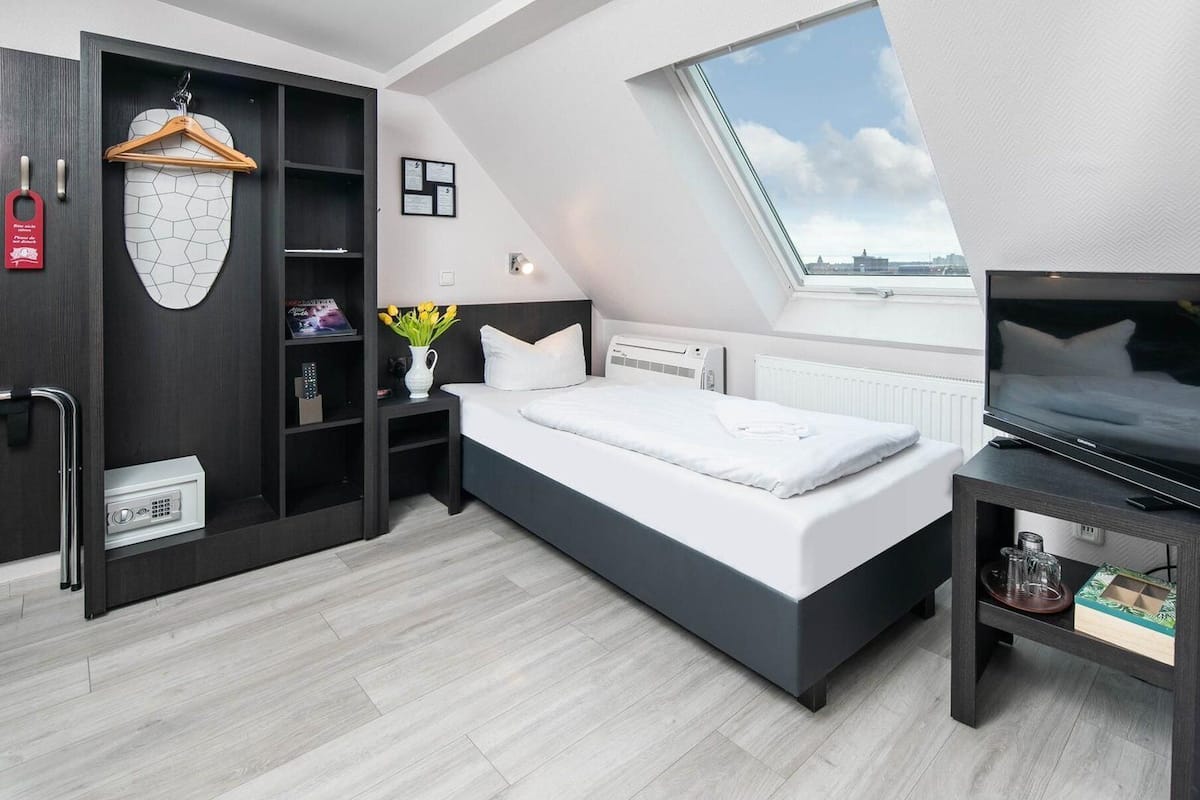 Ein modernes Hotelzimmer mit einem großen Bett, einem Fernseher, einem Schrank mit Spiegel, einem kleinen Tisch mit Blumen und einem Fenster mit Blick auf die Stadt.