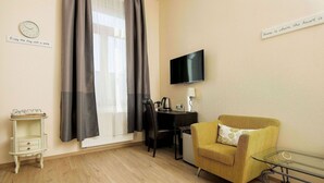 A room with a wooden floor, a yellow armchair, a glass table, a TV, a desk with a lamp, a clock, and a message on the wall. - City Hotel Kaiserhof (Offenbach am Main)