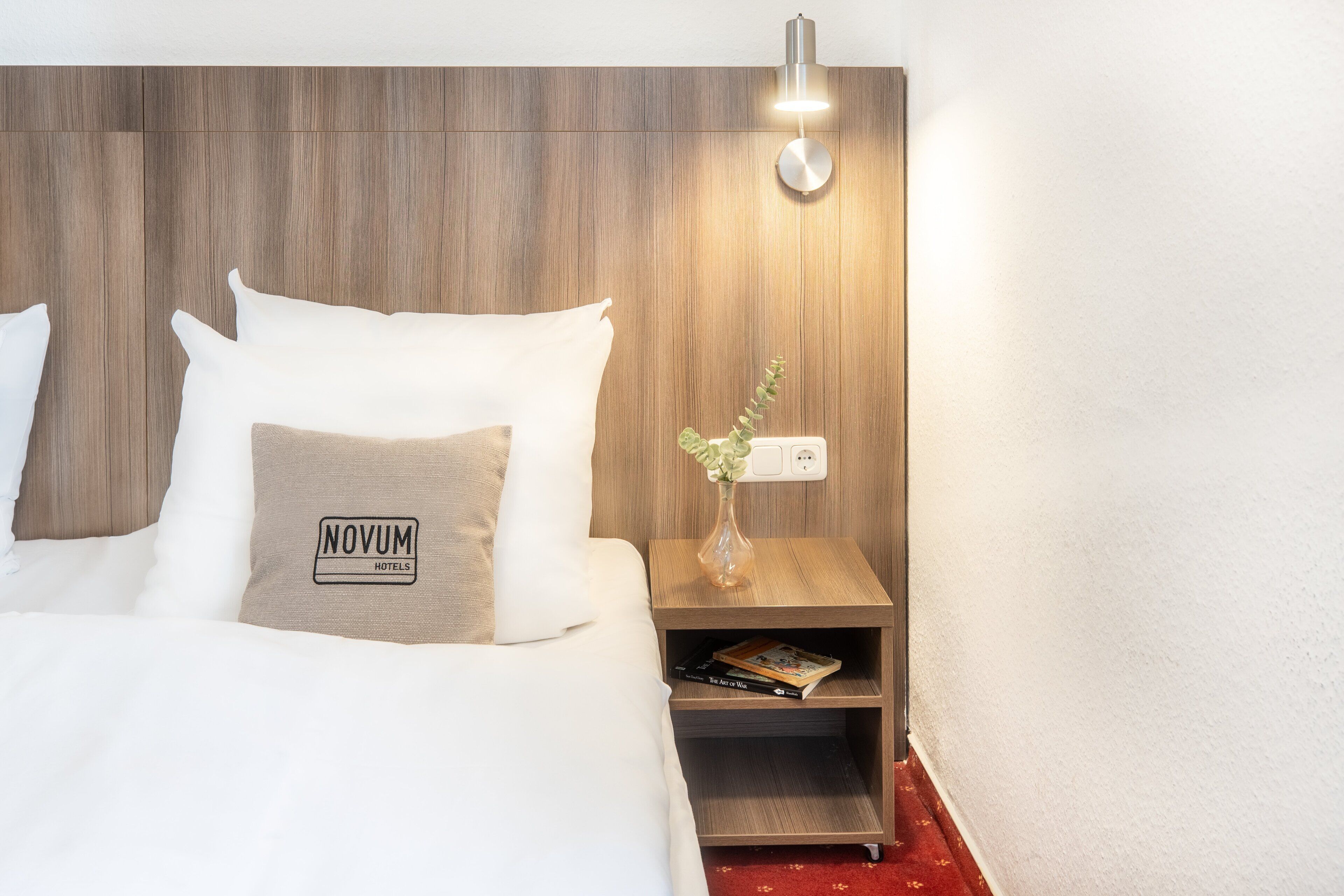 A hotel room with a wooden headboard, a bed with white bedding, a nightstand with a vase of flowers, and a sign reading "NOVUM HOTELS".