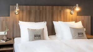 Ein Hotelzimmer mit Holz-Kopfteil, zwei Nachttischen, weißer Bettwäsche und Kissen mit dem Novum Hotels Logo.