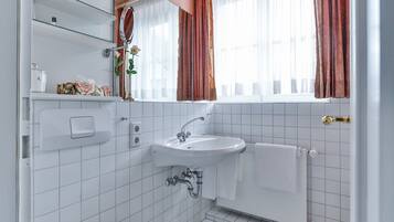 Un baño con lavamanos blanco, espejo y toallero. Se aprecia una ventana con cortinas y un radiador.